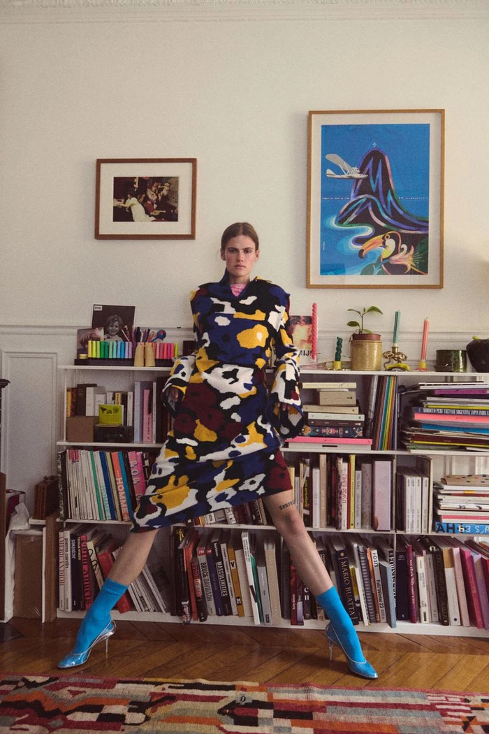 Model standing with wide stance in a colourful patterned coat and bright blue socks against a packed bookshelf