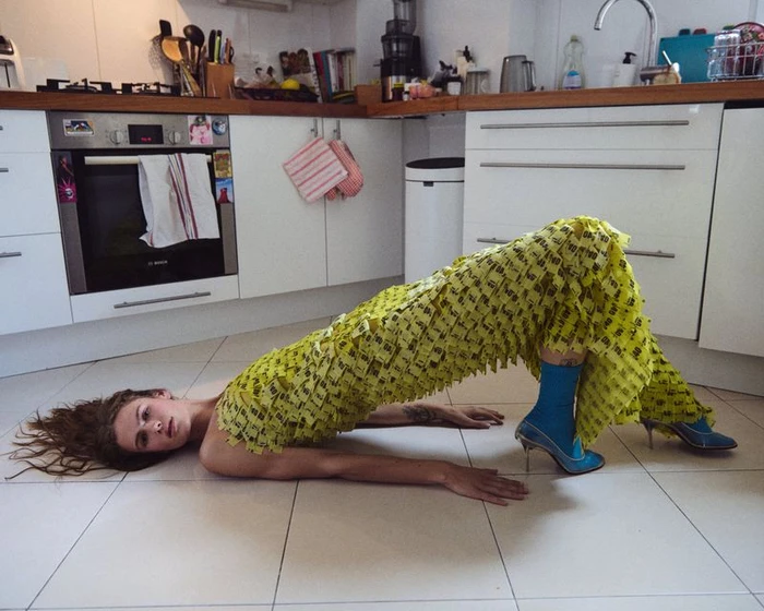 Model lying in a bridge pose on a tiled kitchen floor wearing a textured yellow fringed dress and blue heeled boots