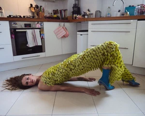 Model lying in a bridge pose on a tiled kitchen floor wearing a textured yellow fringed dress and blue heeled boots