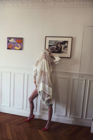 Model draped in white sheer fabric wearing red heels leaning against a paneled wall in an interior setting.