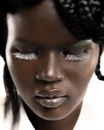 Close-up portrait of a Black woman with braided hair, white eyelashes, and glossy lips against a bright white background.