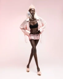 Model in pink feathered hat and sheer oversized shirt posing against a soft pink background