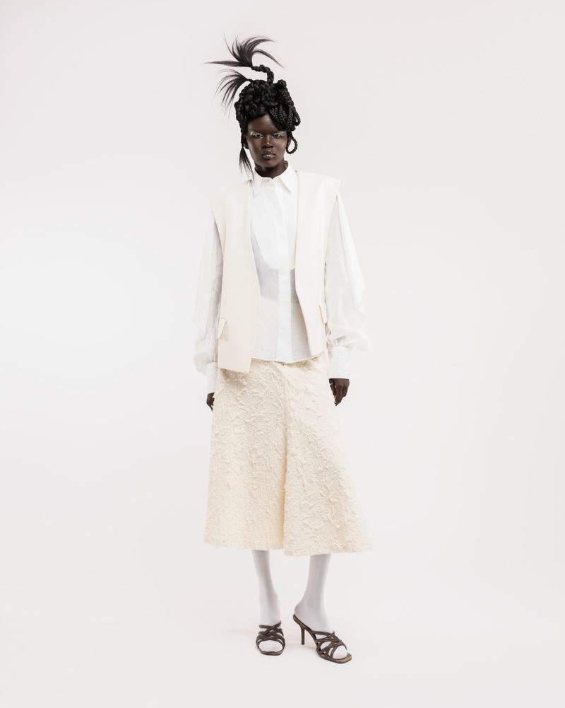 Model wearing a white shirt, beige vest, textured skirt, and structured hairstyle against a plain white background