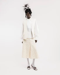 Model wearing a white shirt, beige vest, textured skirt, and structured hairstyle against a plain white background