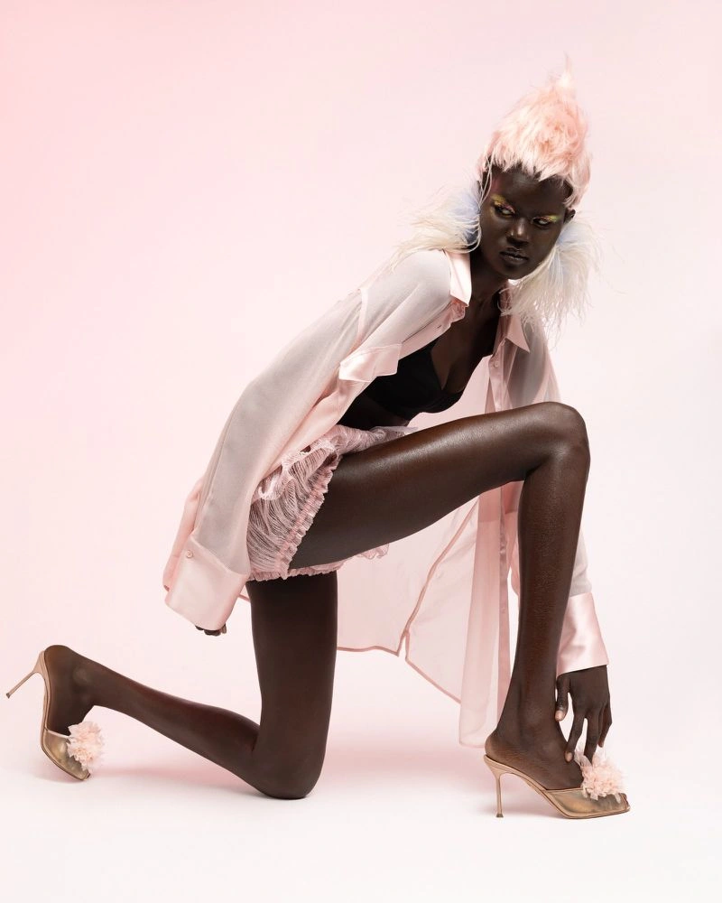 Model kneeling on pale studio backdrop wearing pink sheer shirt, lace shorts, and feathered heels