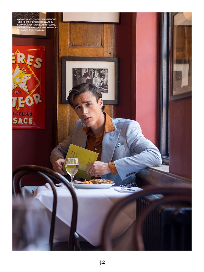 Man in a light grey suit jacket seated at a restaurant table with a glass of white wine, food, and a yellow menu.