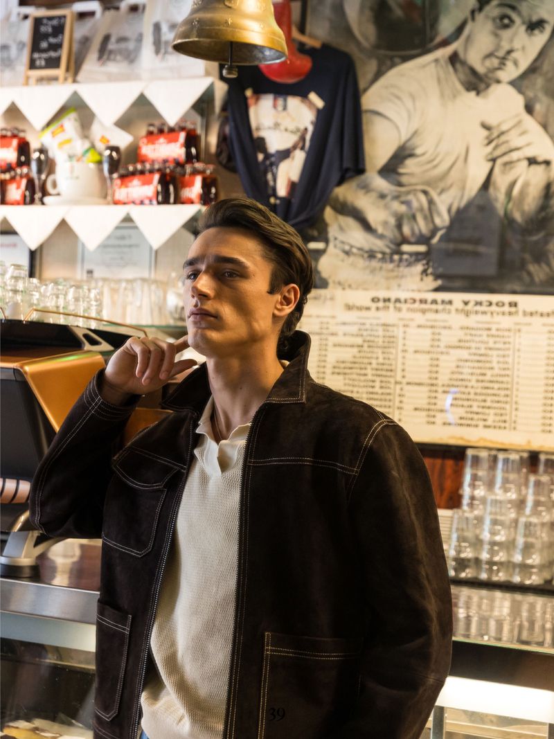 Indoor portrait of a young adult man in a dark brown jacket and white polo shirt in an urban setting with a bell and menu board.