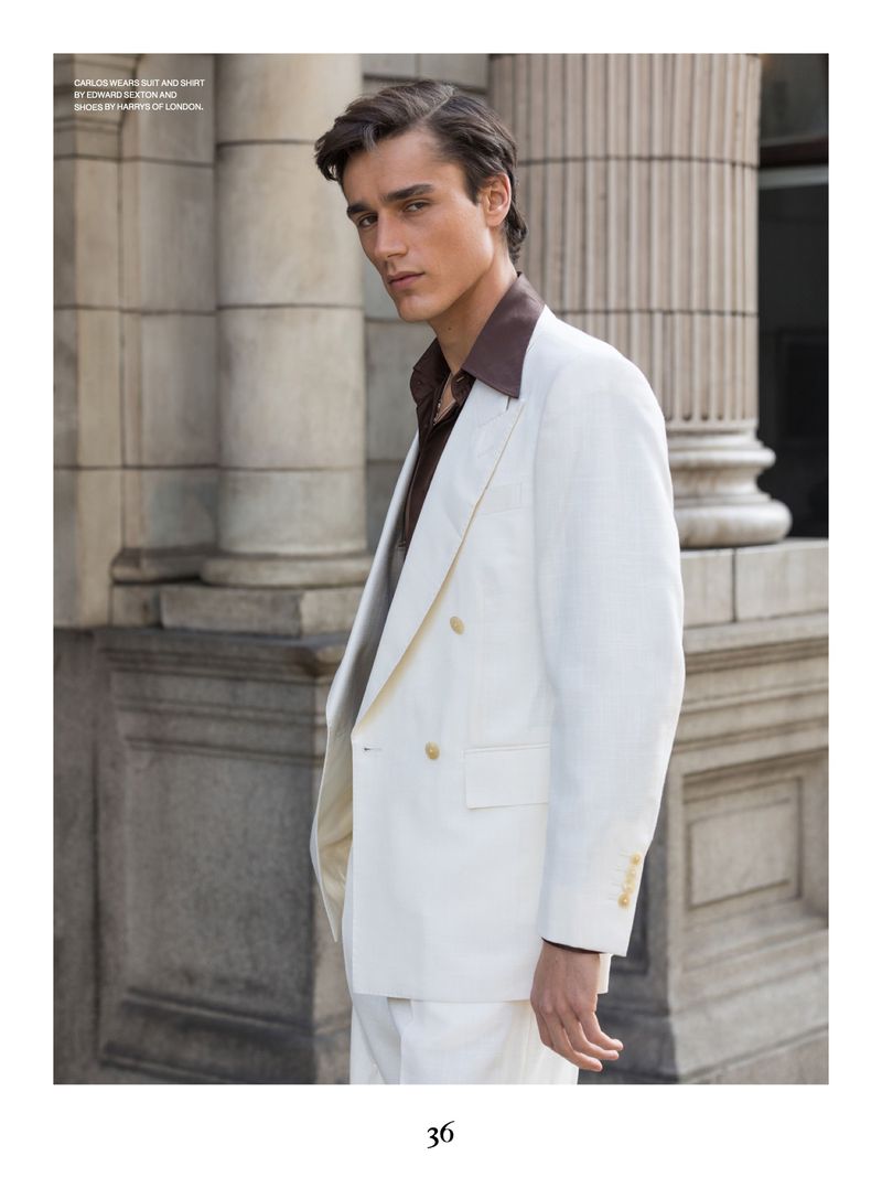 A fair-skinned male model in a white suit and brown shirt stands in front of a columned building, showcasing a blend of classic style.