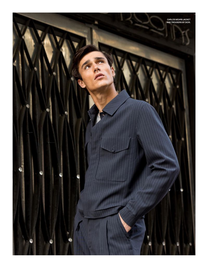 Adult male fashion model poses in a pinstripe jacket and pants in front of a dark metal gate looking up in an outdoor setting.