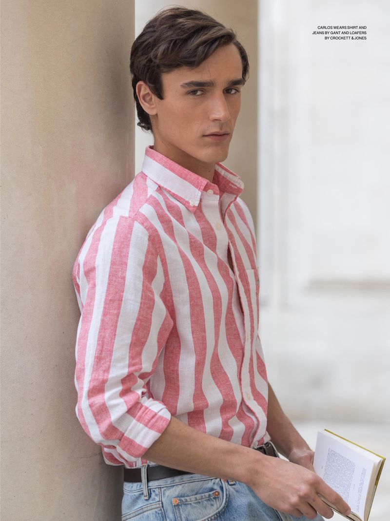 Young adult male in vertical striped button-down shirt and jeans leaning against a wall holding a book in a bright, airy indoor space.