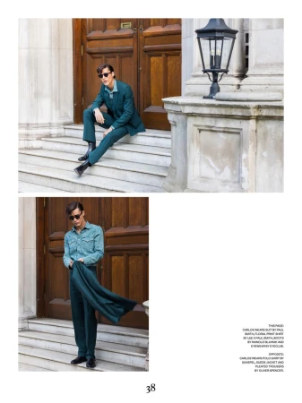 Man in green suit poses on steps of building with wooden door, wearing sunglasses and black boots for outdoor fashion editorial.