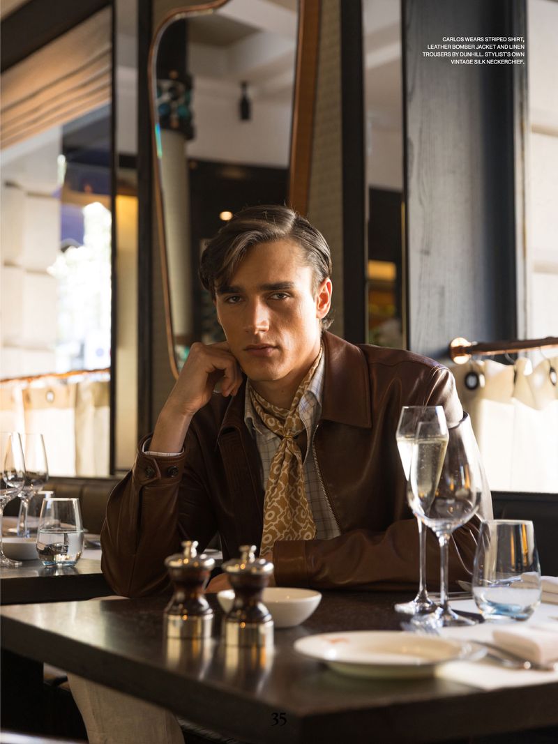 Stylish brunette man in a brown jacket poses in a restaurant setting with champagne glasses, tableware, and elegant decor.