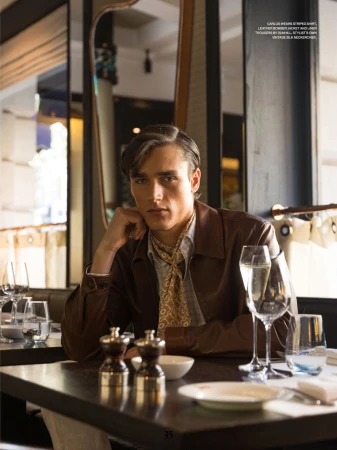Stylish brunette man in a brown jacket poses in a restaurant setting with champagne glasses, tableware, and elegant decor.