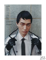 Asian man in white shirt, dotted tie, and clear plastic coat with black leather gloves against a stone wall.