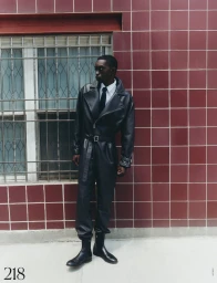 Man wearing a black leather coat and trousers standing against a dark red tiled wall next to a barred window