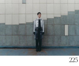 Model standing against a tiled exterior wall wearing a clear vinyl coat, white shirt, dark tie, and pinstriped trousers.