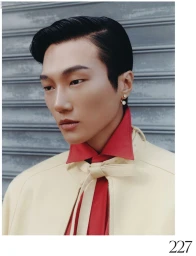 Close-up portrait of an East Asian male model wearing a red collared shirt and cream jacket against a ribbed metal shutter.