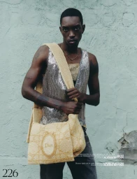 Black man holding a large woven straw shoulder bag wearing a silver sequined tank top against a textured turquoise wall