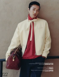 Asian man in cream jacket and red shirt leaning against textured wall holding a dark red leather duffel bag.