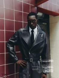 Black man wearing a black leather coat with a white shirt and black tie posing against a red tiled wall.