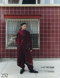 Model in burgundy layered coat and wide trousers standing before dark red tiled wall and barred window for French Magazine.