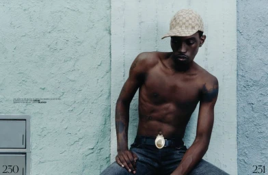 Shirtless man wearing monogram print cap and dark jeans posing outdoors against a textured light blue and white wall.