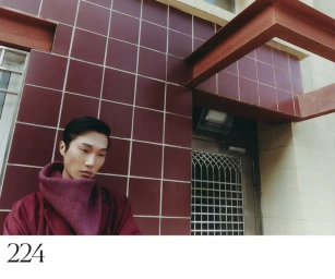 Model wearing a structured burgundy coat standing against a dark red tiled building exterior with architectural elements