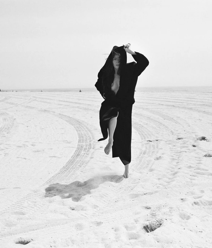 Havana Rose Liu in a black hooded coat leaping across a sandy beach for Flaunt magazine, photographed by Arno Frugier.