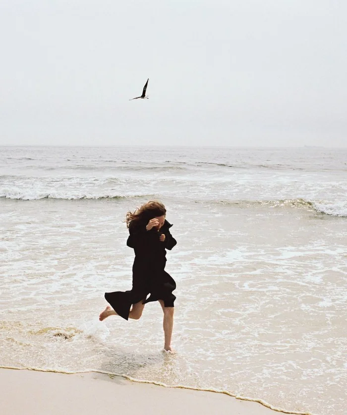 Havana Rose Liu running barefoot in the surf along a sandy beach under an overcast sky as a bird flies overhead.