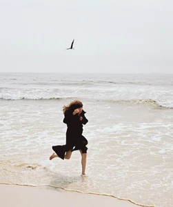 Havana Rose Liu running barefoot in the surf along a sandy beach under an overcast sky as a bird flies overhead.