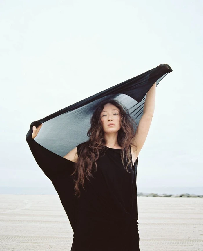 Havana Rose Liu holding a black fabric above her head against an open sandy beach and pale sky background