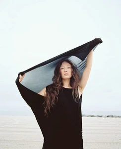 Havana Rose Liu holding a black fabric above her head against an open sandy beach and pale sky background