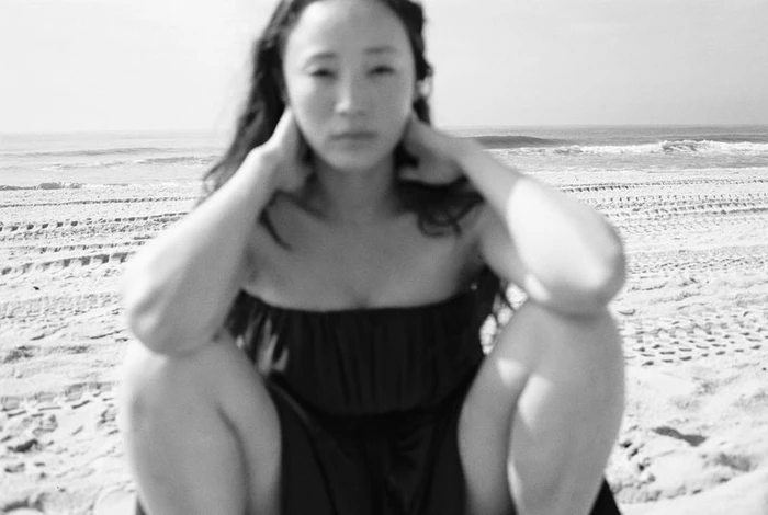 Havana Rose Liu squatting on a sandy beach with the ocean behind her in a black strapless dress captured by Arno Frugier for Flaunt