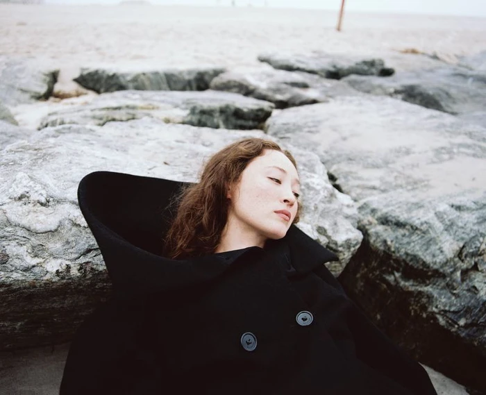 Havana Rose Liu reclining among coastal rocks wearing a structured black coat with a high collar, photographed by Arno Frugier.