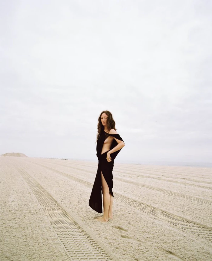 Havana Rose Liu wearing a black dress standing on a sandy beach with tire tracks under an overcast sky.