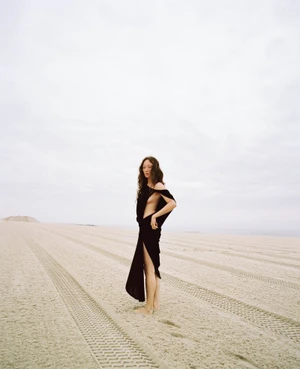 Havana Rose Liu wearing a black dress standing on a sandy beach with tire tracks under an overcast sky.