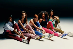 Five female models in athletic wear seated in a row against a dark studio background for V Magazine.