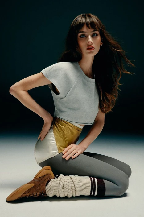 Model with dark hair wearing a grey crop top and metallic leggings kneeling in a studio setting.