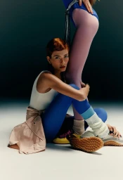 Model with red hair seated on the floor wearing a white tank top and blue leggings with leg warmers, posed against another standing model.