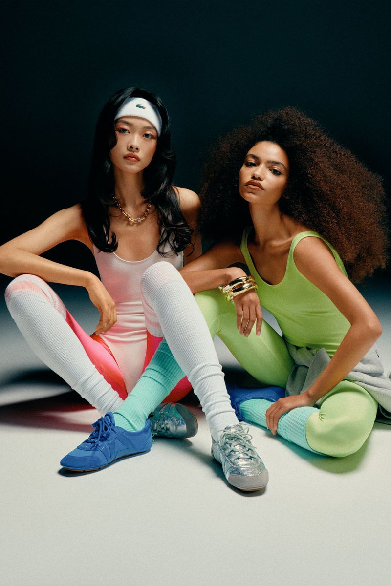 Two female models in athletic wear sitting against a dark gradient studio background for V Magazine.