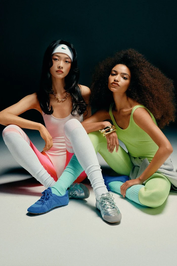 Two female models in athletic wear sitting against a dark gradient studio background for V Magazine.
