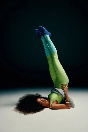 Model in lime green bodysuit and blue sneakers holding a shoulder stand pose against a dark background