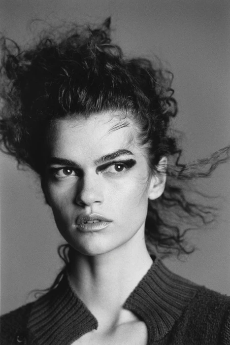 Black and white close-up portrait of a woman with dark curly hair and bold eye makeup against a seamless grey studio backdrop