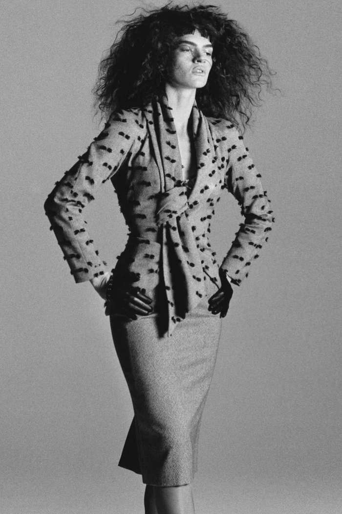 Model with voluminous dark curly hair wearing a patterned jacket and midi skirt against a grey background.