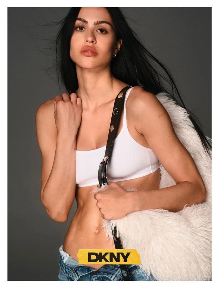 Amelia Gray in a white top and denim jeans holding a fluffy white bag against a grey seamless backdrop