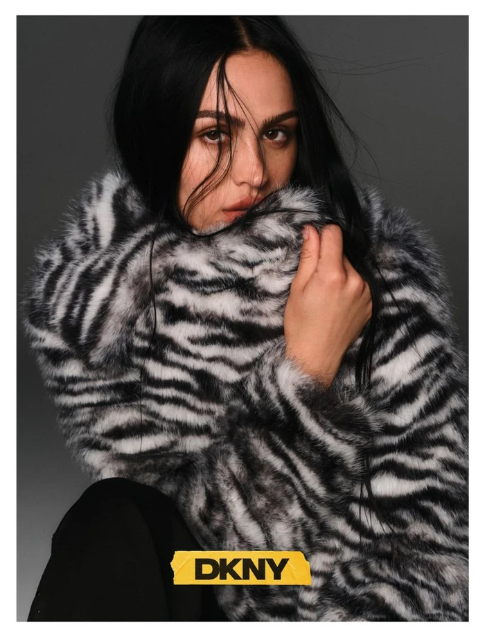 Model Amelia Gray with dark hair wearing a black and white faux fur zebra coat against a grey backdrop for DKNY.