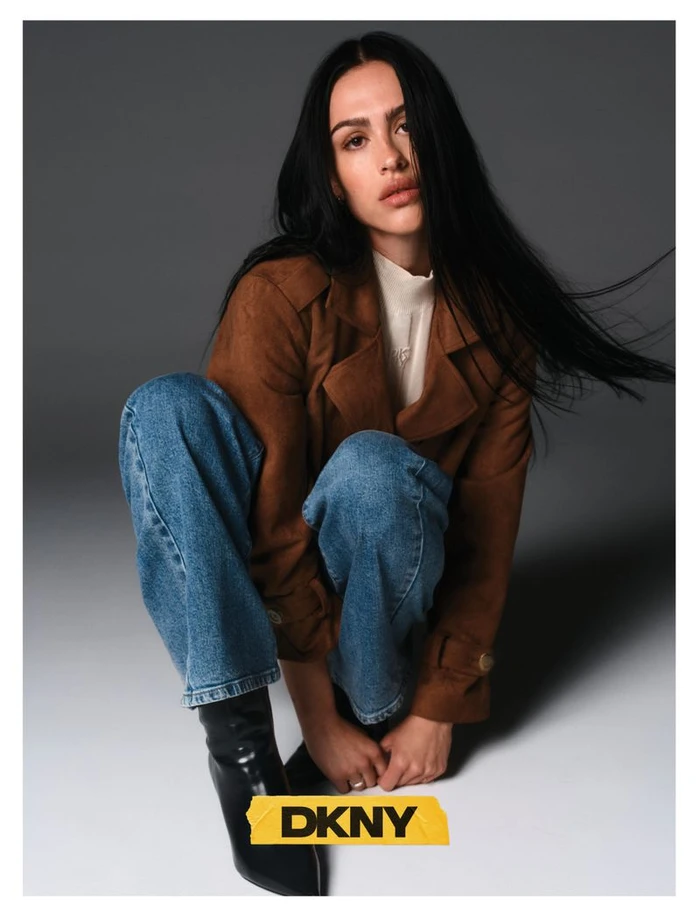 Model Amelia Gray in brown jacket and blue jeans crouching against a grey gradient background for DKNY fall campaign