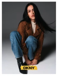 Model Amelia Gray in brown jacket and blue jeans crouching against a grey gradient background for DKNY fall campaign