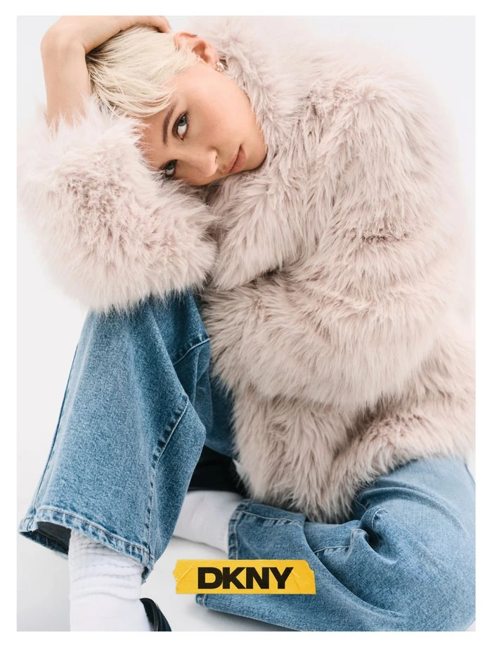 Model with blonde hair wearing a fluffy cream faux fur coat and blue jeans against a white background for DKNY Fall 26 edit.