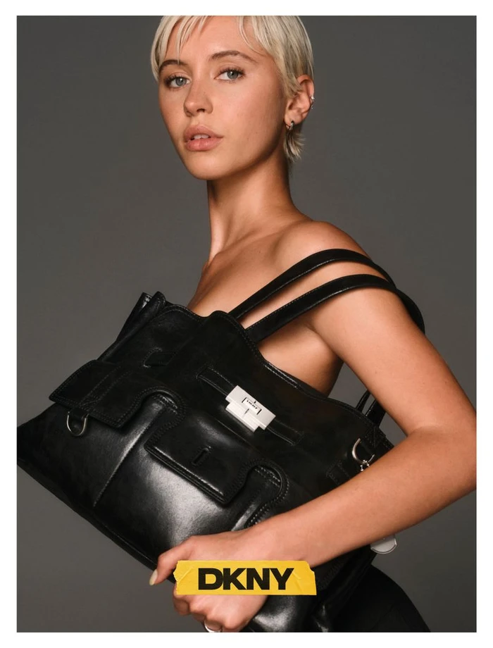 Model Iris Law holding a large black leather handbag against a solid grey studio background for DKNY Fall 26 edit